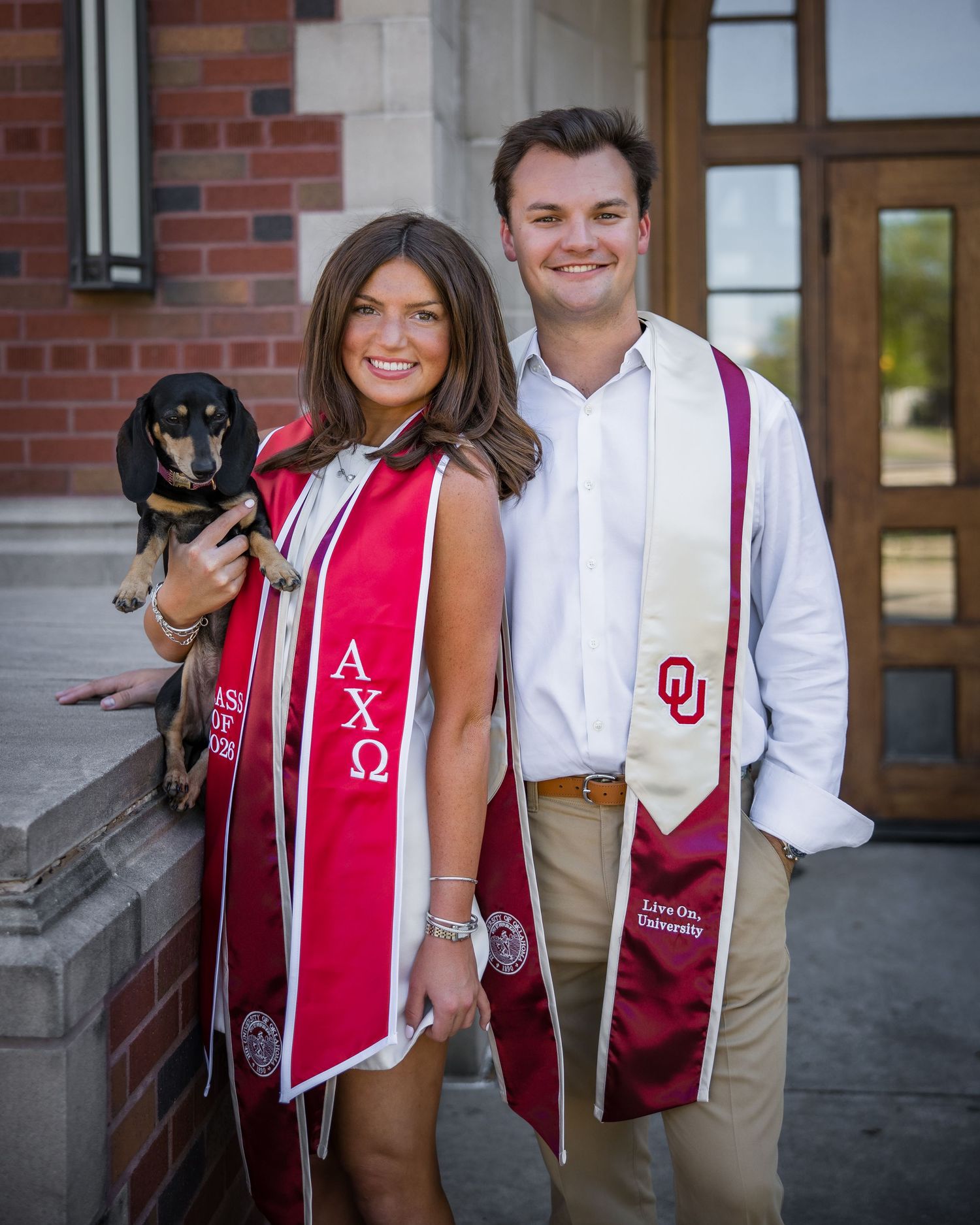 Two University of Oklahoma graduates in red and white stoles smile with a dachshund outside a brick building.