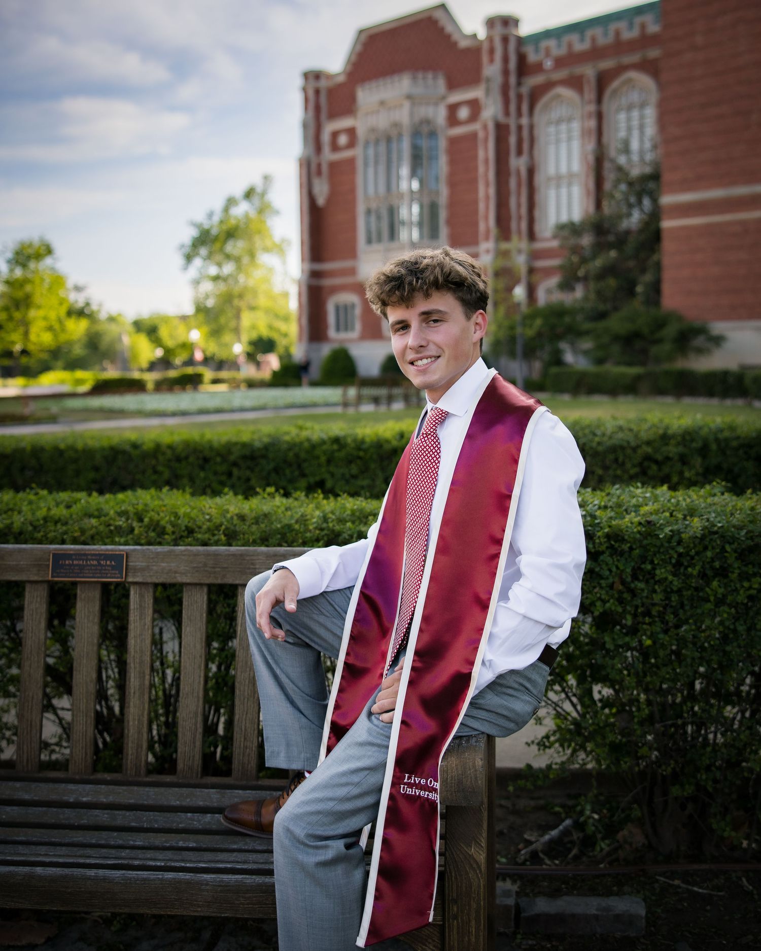 Young man in white shirt and maroon graduation stole sits on bench outside brick university building.