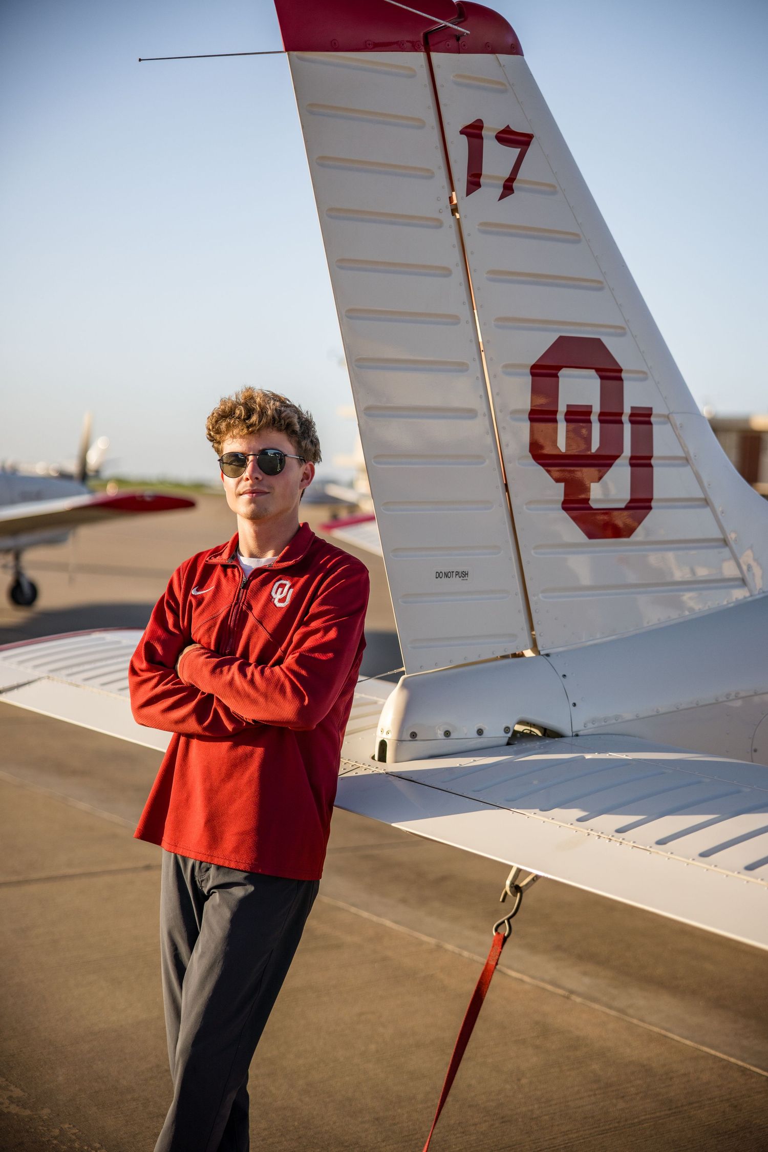 Young man in OU red sweatshirt stands arms crossed beside University of Oklahoma branded airplane tail on tarmac.