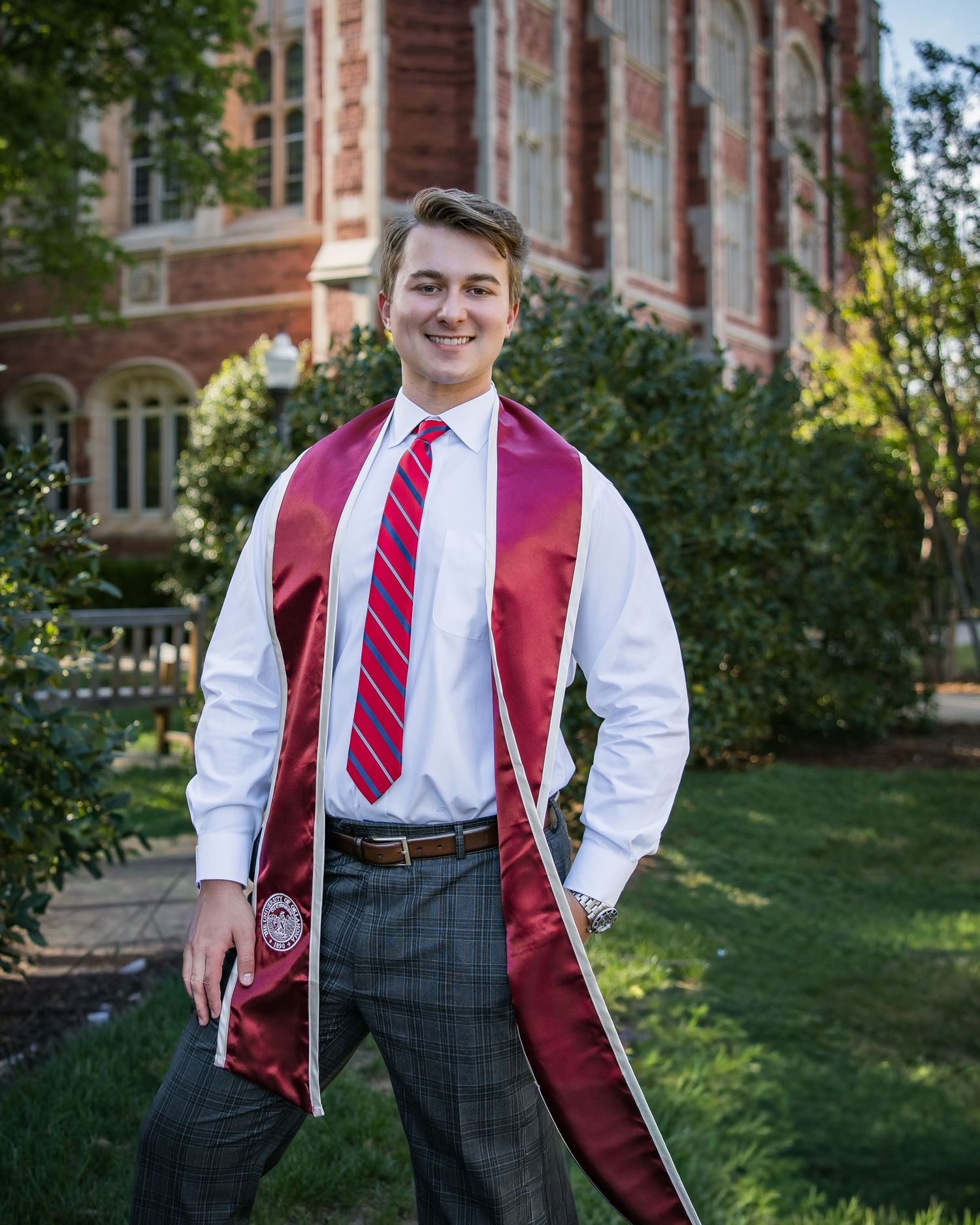Young man in white dress shirt, red tie, and maroon graduation stole smiling outdoors near brick building.