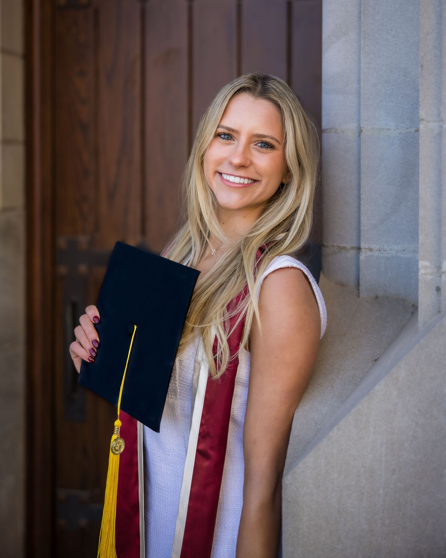 Graduate in burgundy and white regalia holds graduation cap and tassel while posing against stone column.