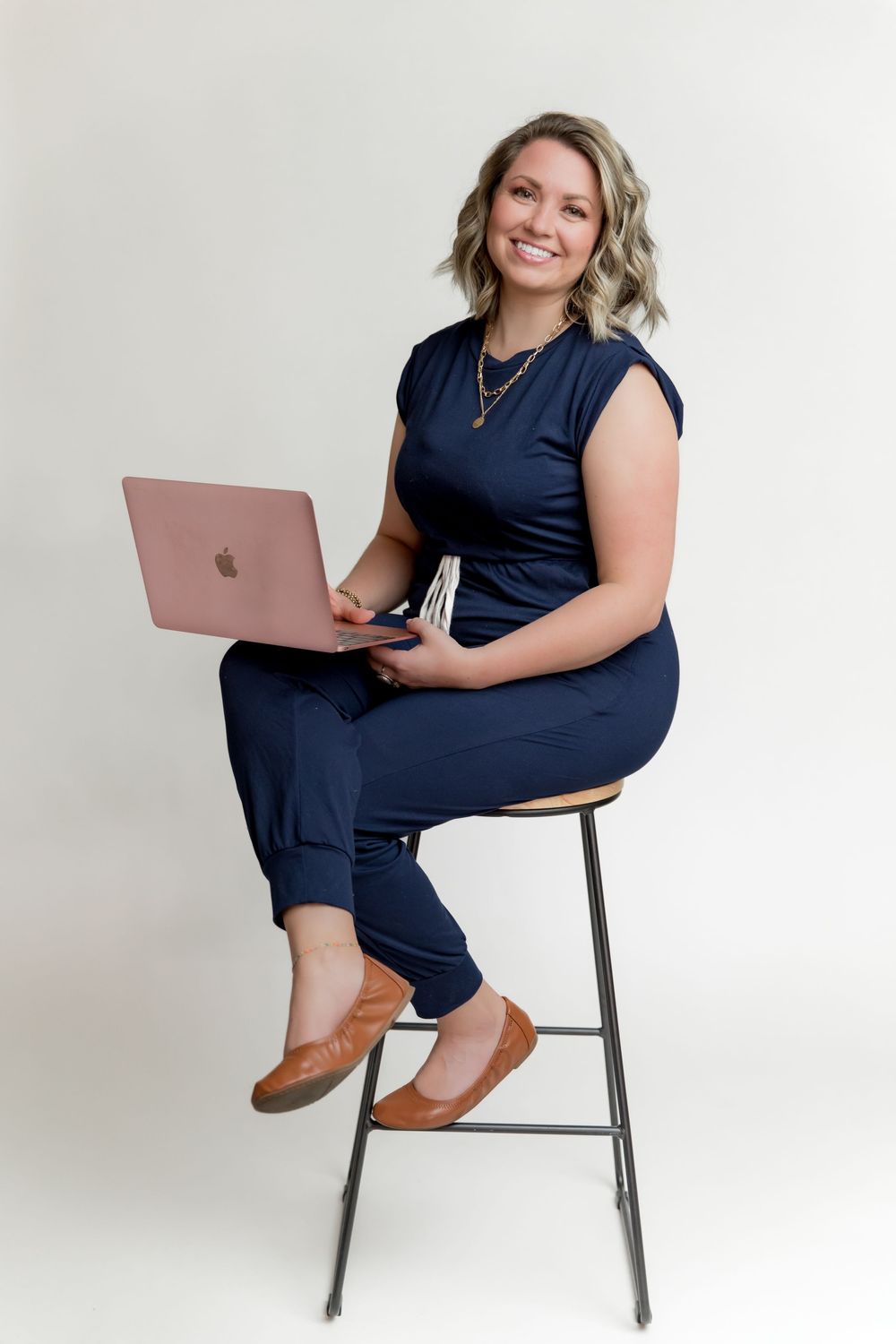 Professional seated in navy dress and brown boots poses with laptop against white backdrop in studio setting.