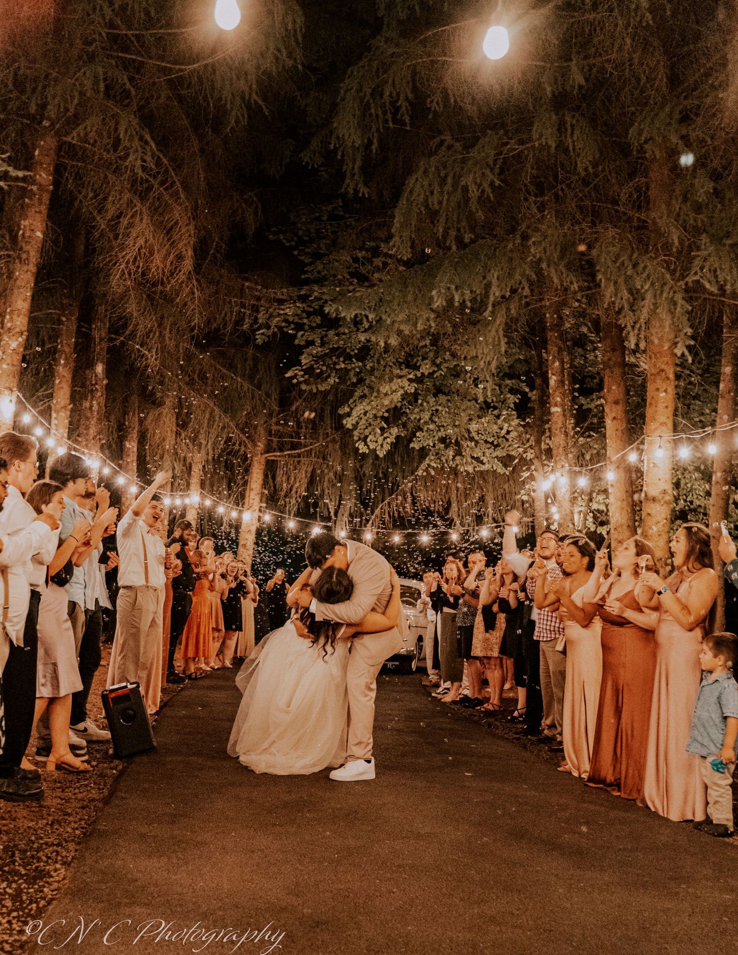 A romantic golden-lit wedding reception with guests lining a tree-lined pathway as newlyweds share a dance under string lights.