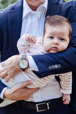 Dad holding his little baby girl. She is looking straight to the camera. Dad is wearing a suite and a beautiful watch.