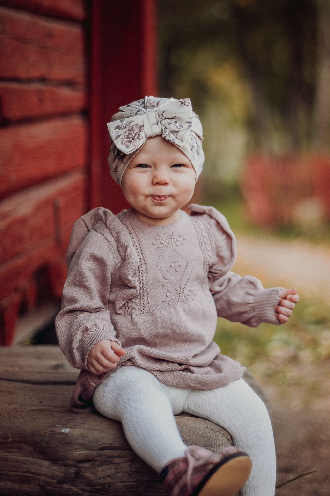 Baby wearing a mauve sweater and white headband sits on wooden surface outdoors.