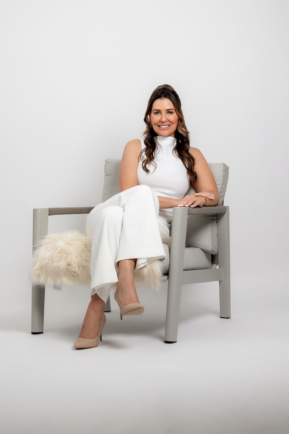 Professional studio photo of a woman in a white dress sitting casually in a modern gray chair against a light backdrop.