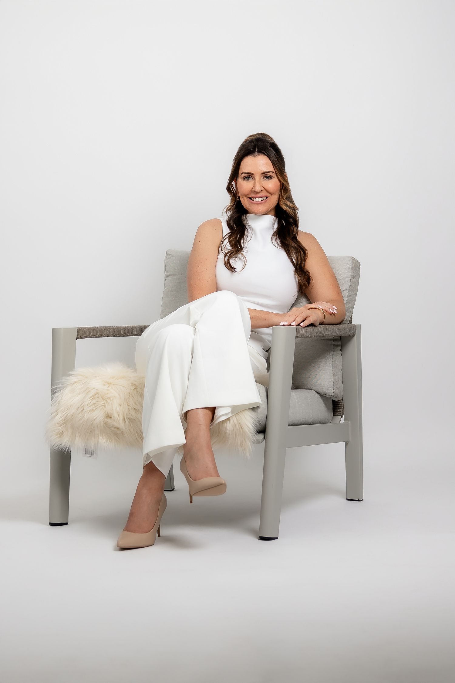 Professional studio photo of a woman in a white dress sitting casually in a modern gray chair against a light backdrop.