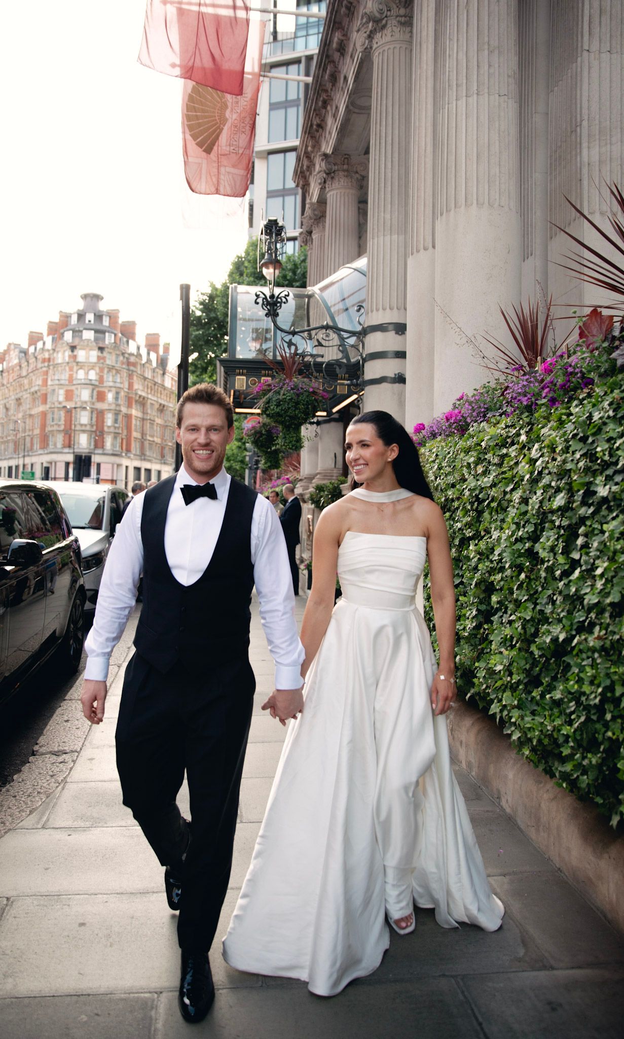 A couple walks hand in hand along a London street, with the bride in a white gown and groom in formal attire.