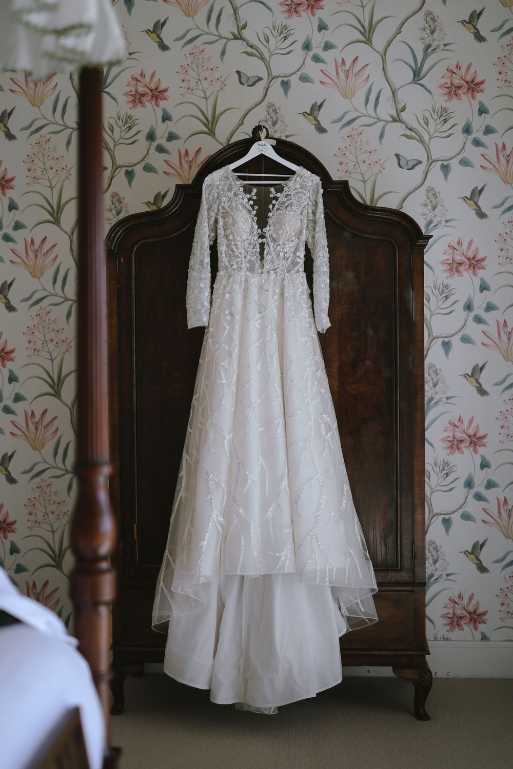 Elegant lace wedding dress with long sleeves hanging on antique wardrobe against floral wallpaper.