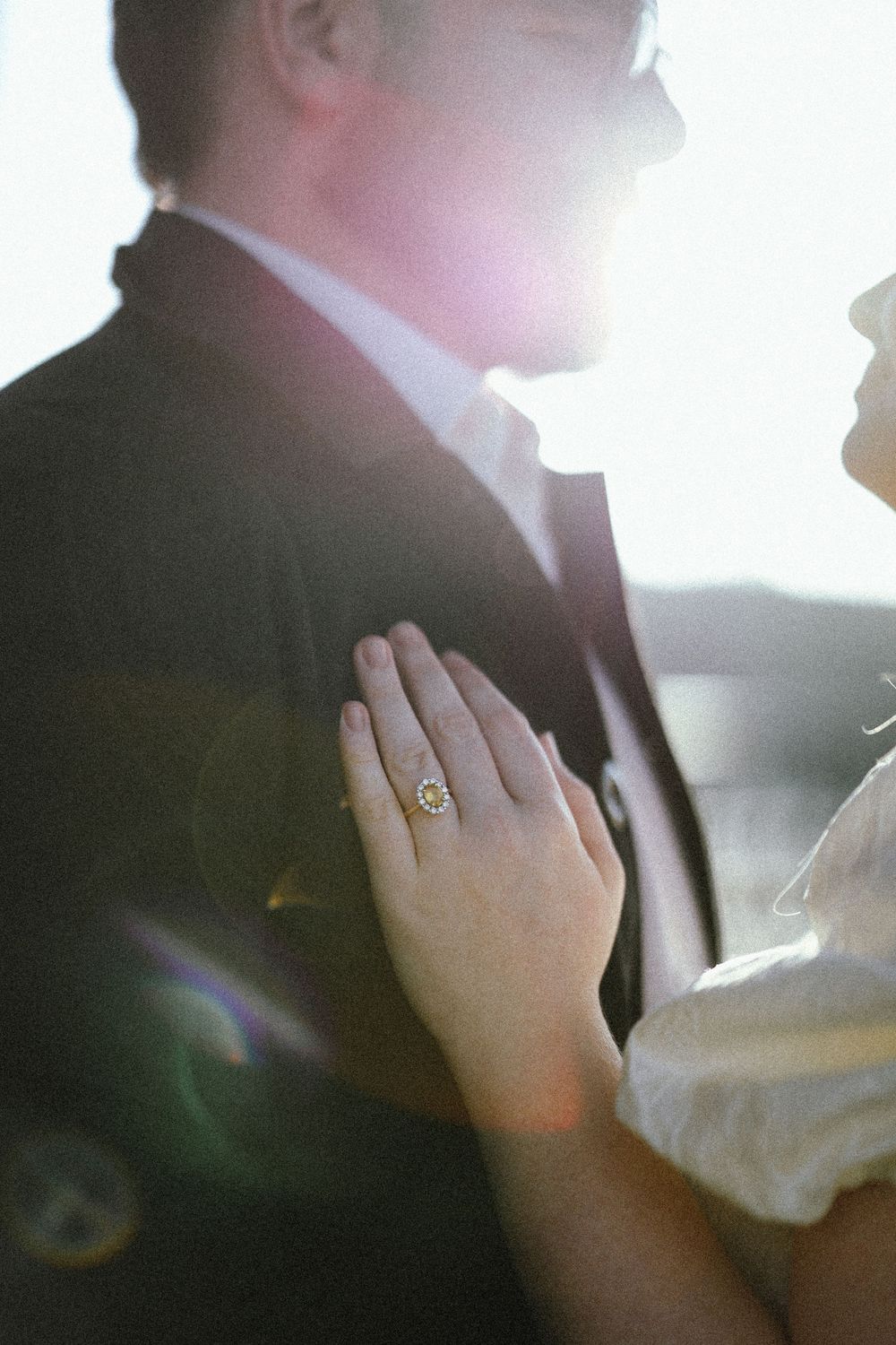Bride resting hand on groom's chest displaying elegant gold engagement ring, backlit by soft sunlight.