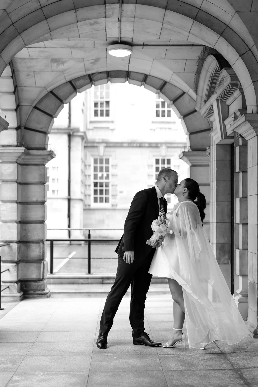 Black and white photo of a bride and groom sharing a kiss under a stone arch at an elegant wedding venue.