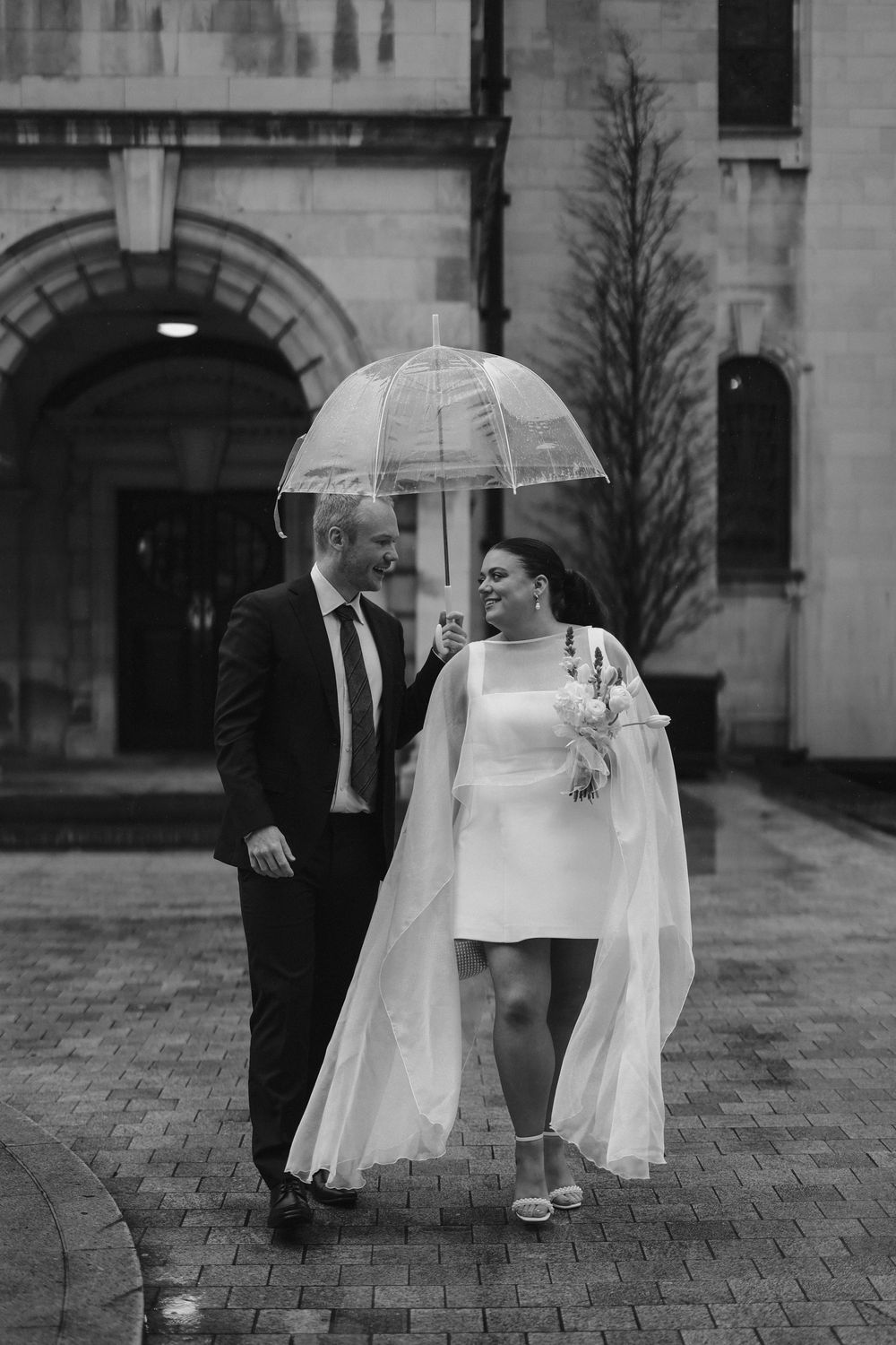 Black and white photo of a bride and groom walking under a clear umbrella on a rainy wedding day.