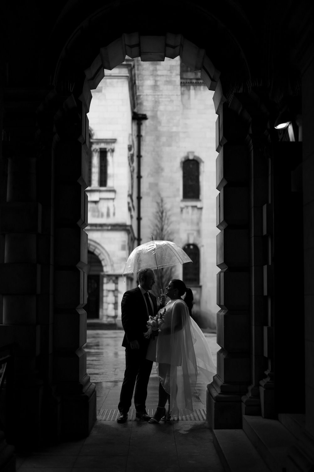 Black and white wedding photo of bride and groom sharing umbrella under stone archway in the rain.