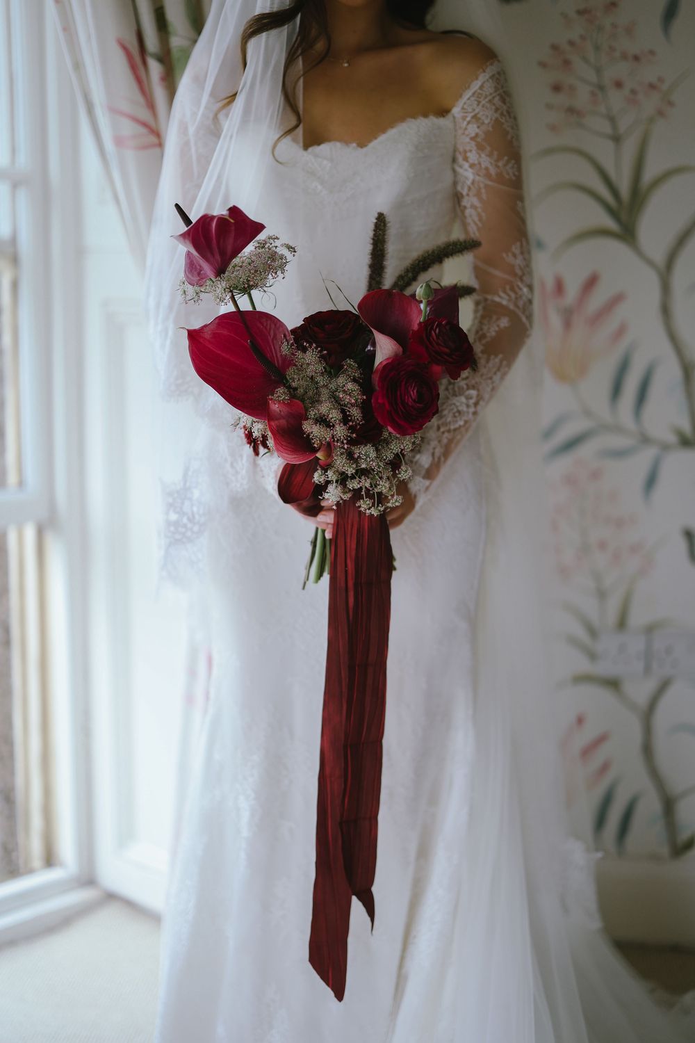 Bride in lace wedding dress holding deep red bouquet with roses, anthuriums, and long burgundy ribbon streamers.