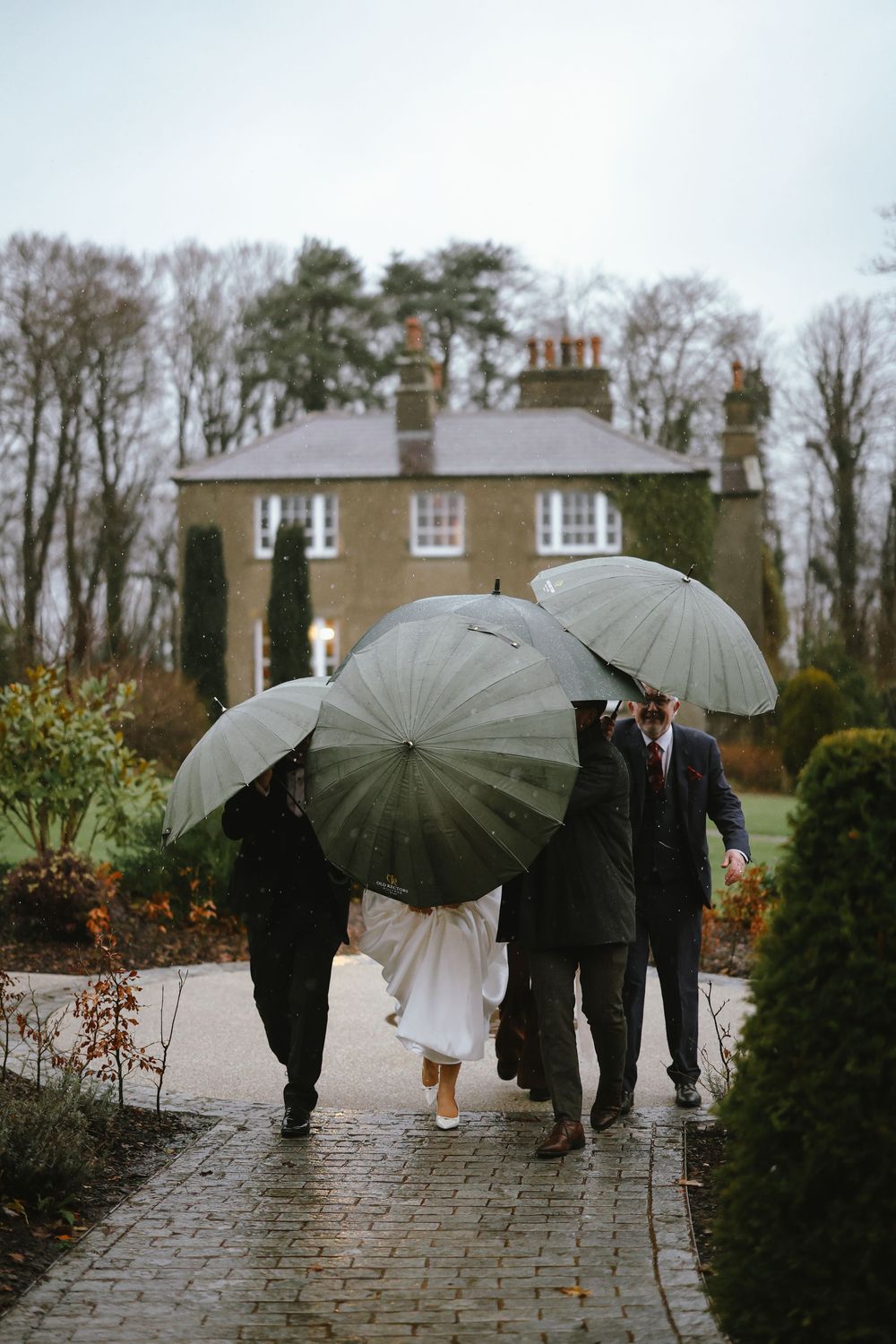 Three people walking with clear umbrellas on a rainy day toward a stone country house surrounded by autumn trees.
