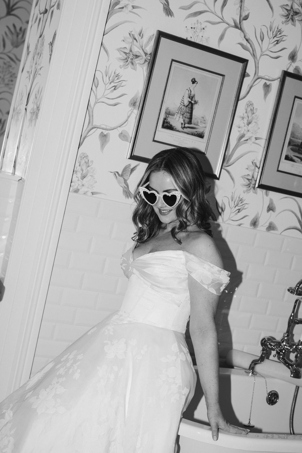 Bride in white off-shoulder gown wearing heart-shaped sunglasses poses in vintage bathroom, black and white photo.