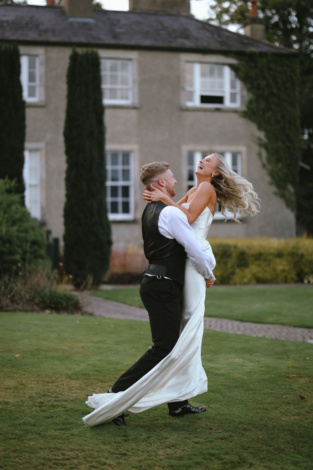 A couple in wedding attire perform a dramatic romantic dip on a lush green lawn at a stately manor house.