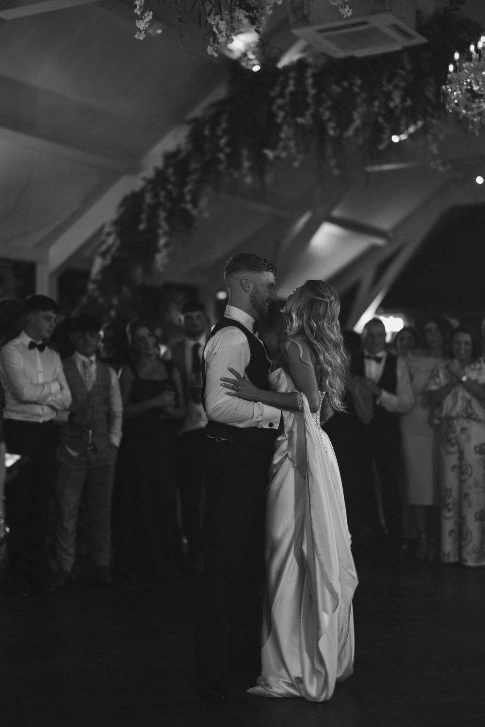Black and white photo of bride and groom sharing first dance at wedding reception with guests watching.