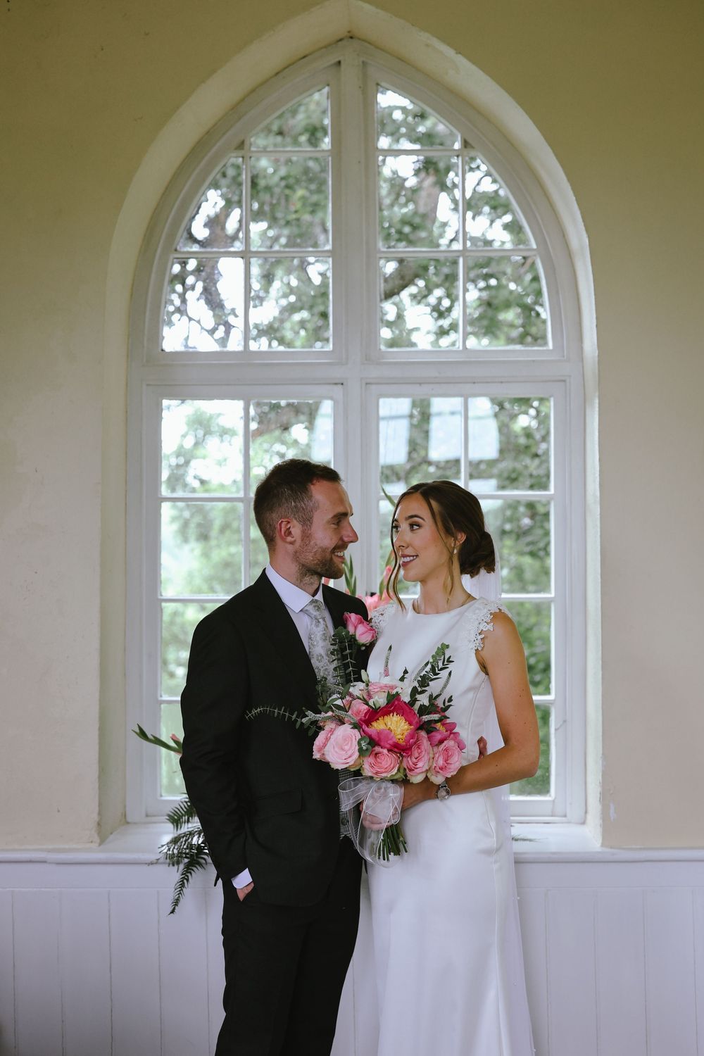 A couple shares a joyful moment together in front of an arched window during their wedding day celebration.