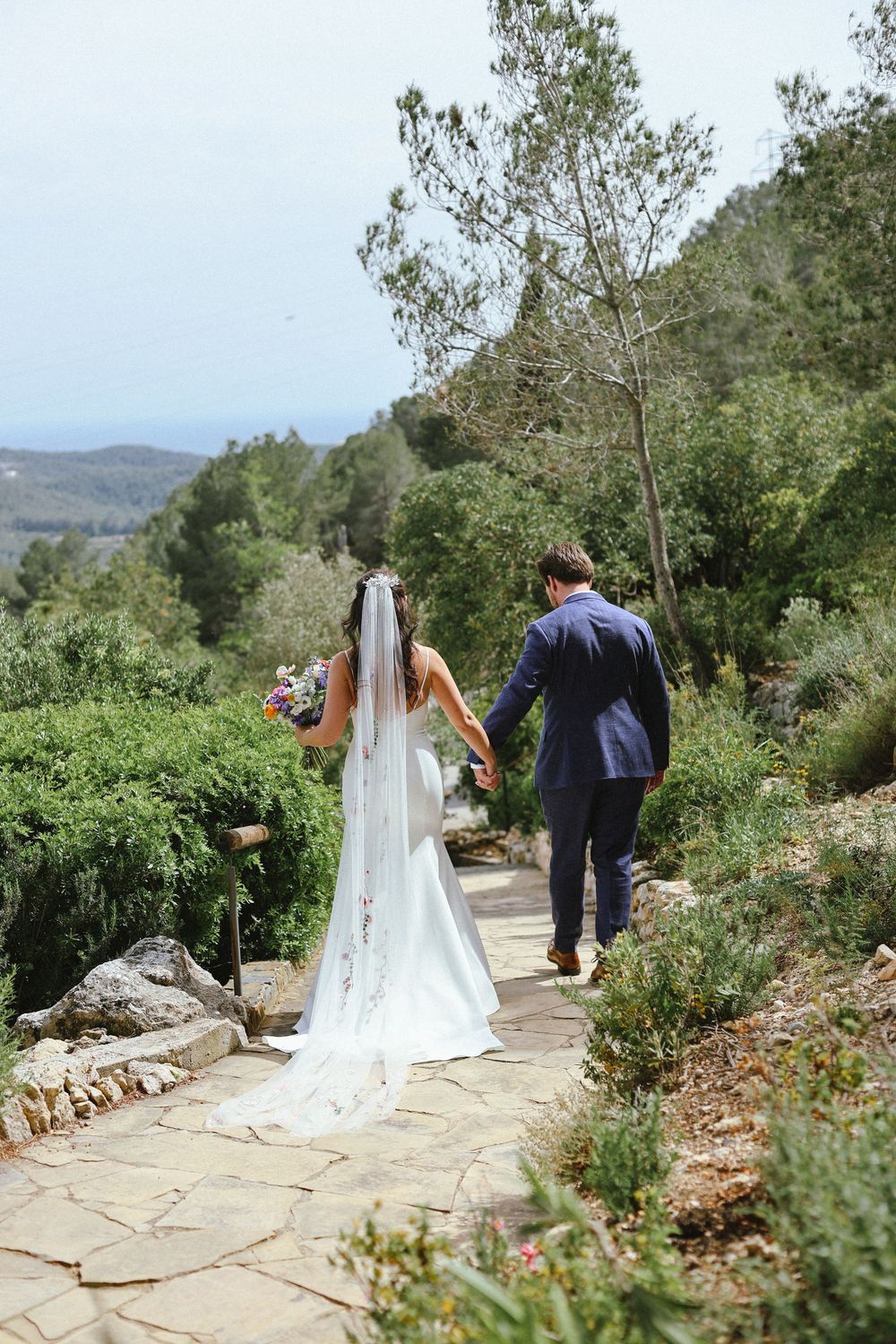 Newlyweds walking hand in hand along a scenic stone path surrounded by lush greenery and mountain views.