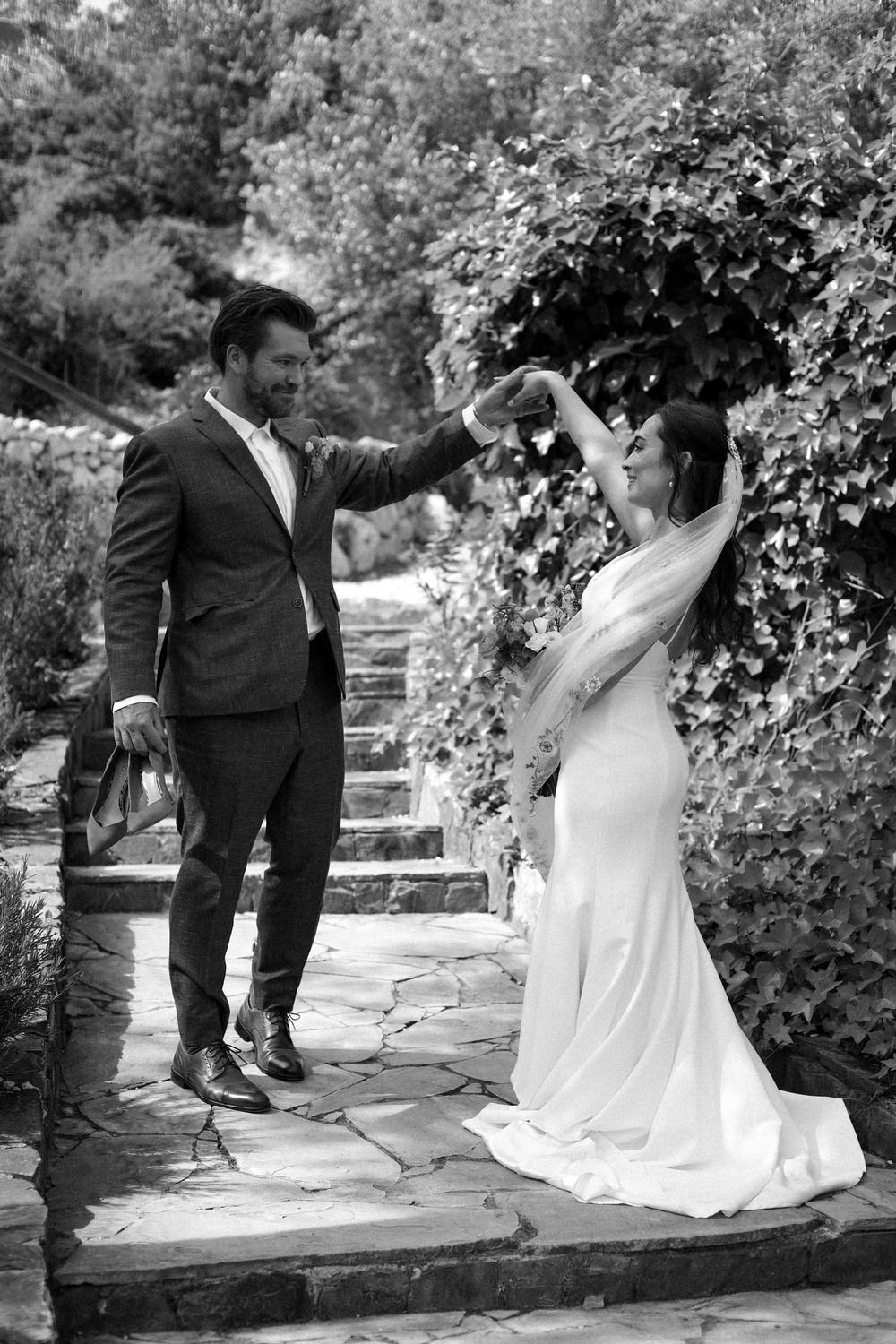 A couple shares a romantic moment in a garden walkway during their outdoor wedding celebration.