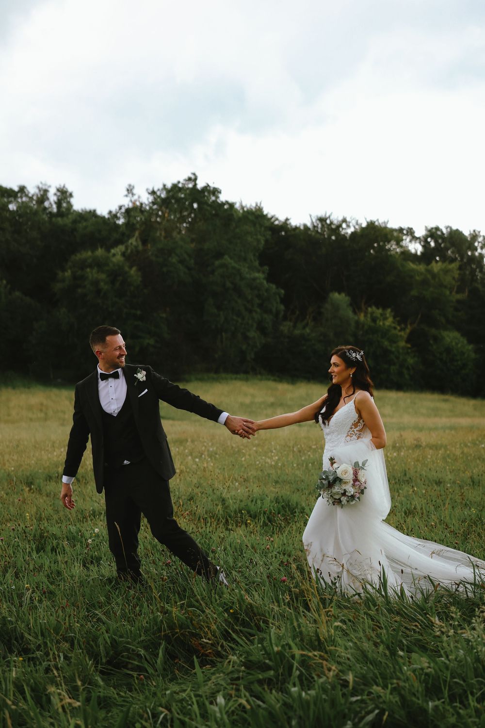 A couple in wedding attire walks through a grassy meadow at sunset, holding hands against a backdrop of trees.