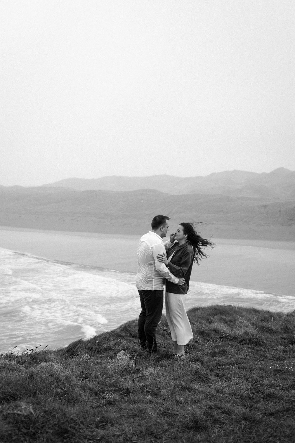 A couple shares a romantic moment on a scenic clifftop overlooking a dramatic coastline in black and white.
