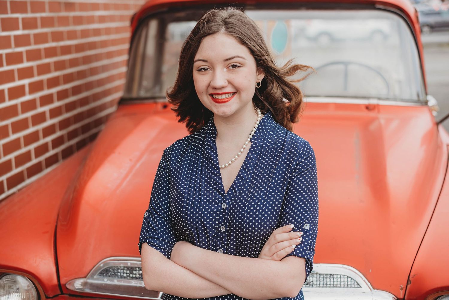 Woman in polka dot dress stands with arms crossed in front of vintage orange car against brick wall.