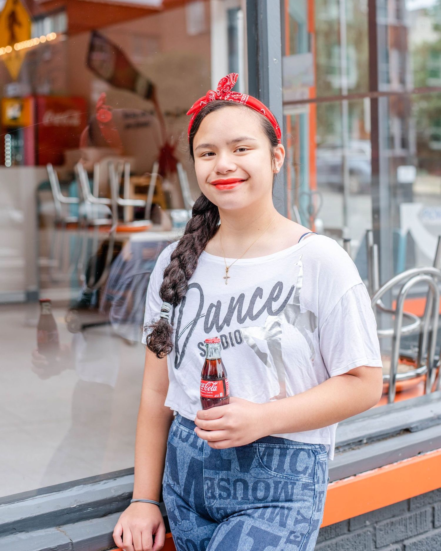 Person in white t-shirt and denim overalls poses with Coca-Cola bottle at retro storefront.