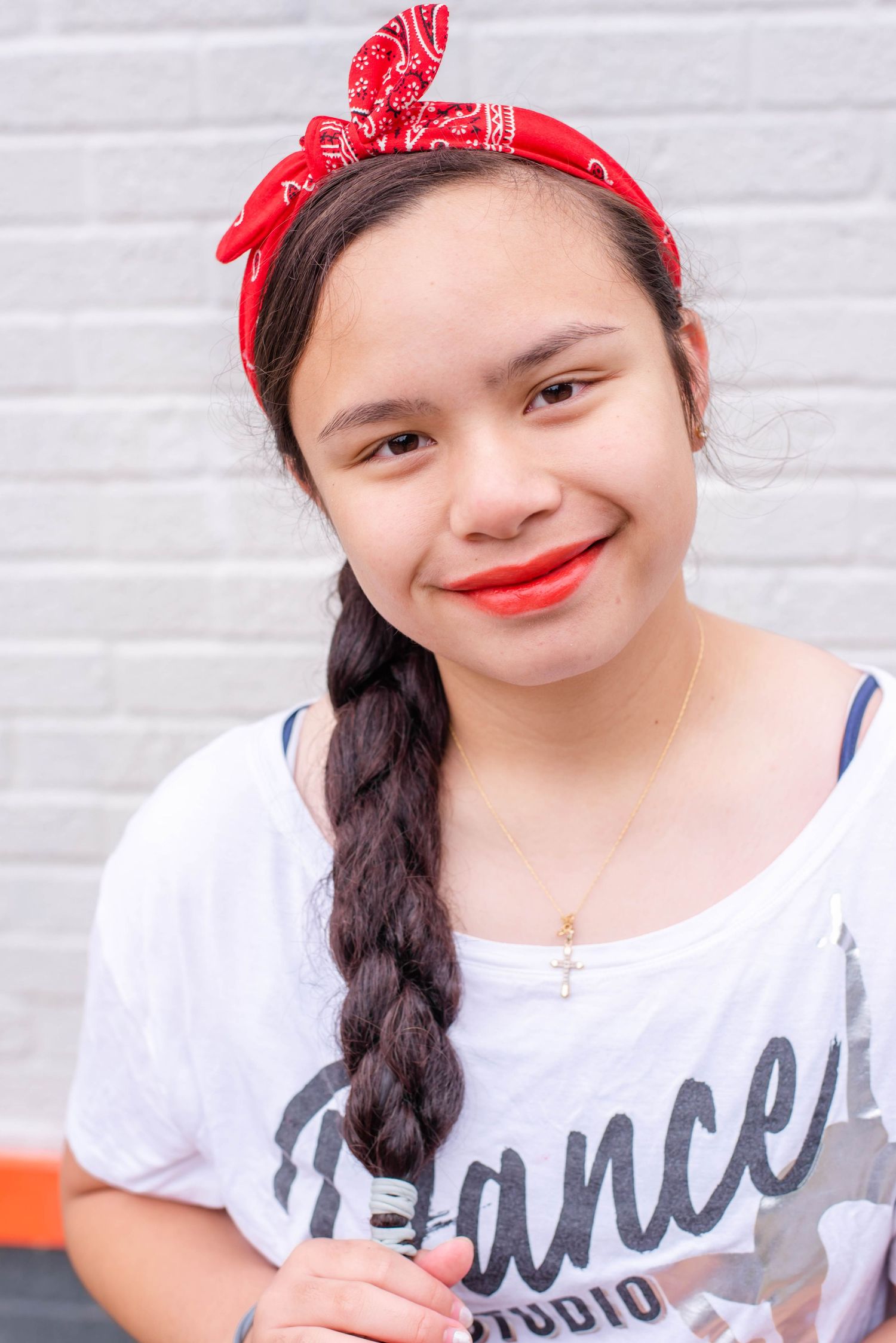 Person wearing a red bandana and white graphic t-shirt smiles at camera in front of white brick wall.