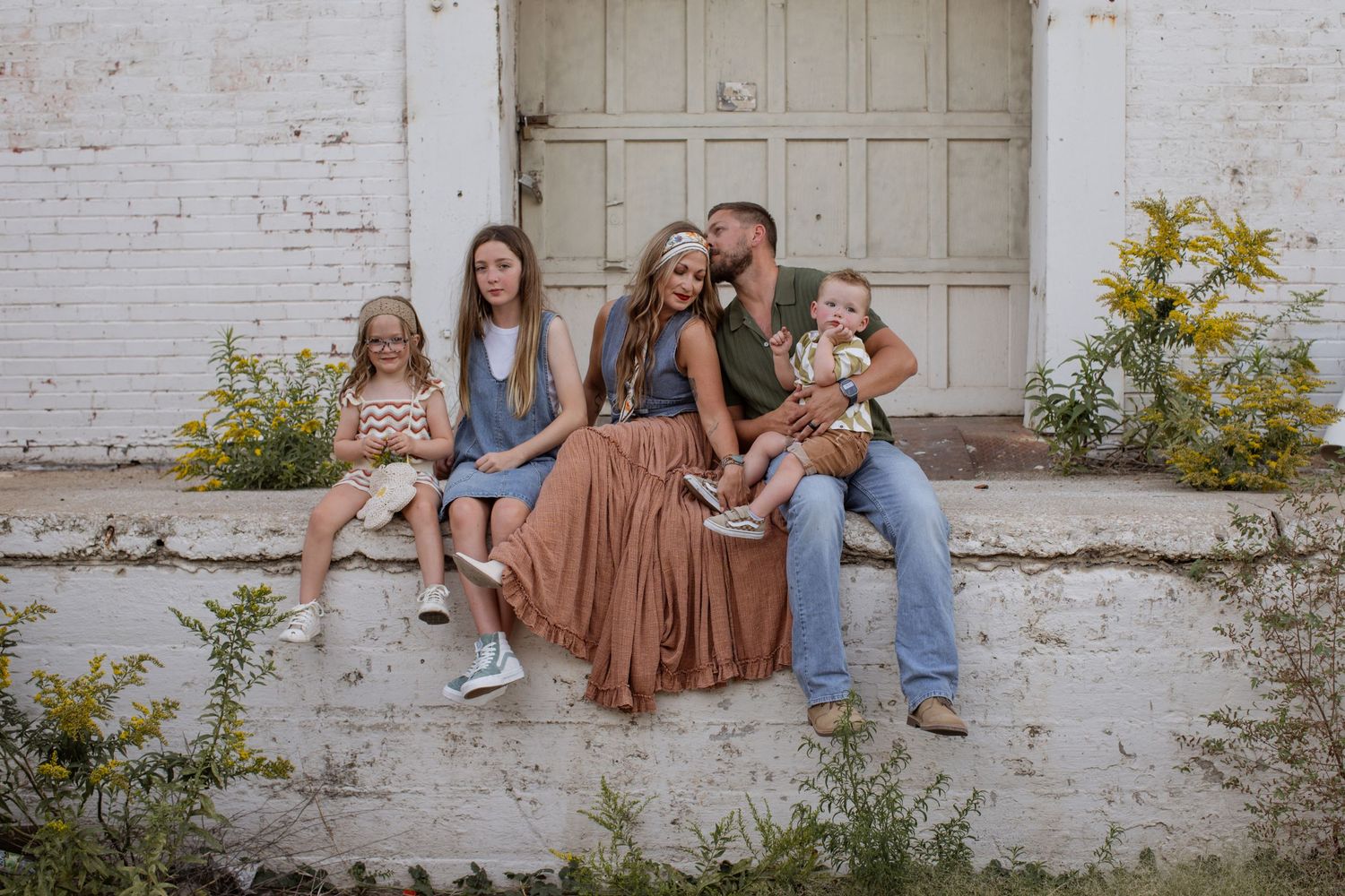A family of five sits on a white-washed wall surrounded by yellow wildflowers during a Kansas City family photo session.