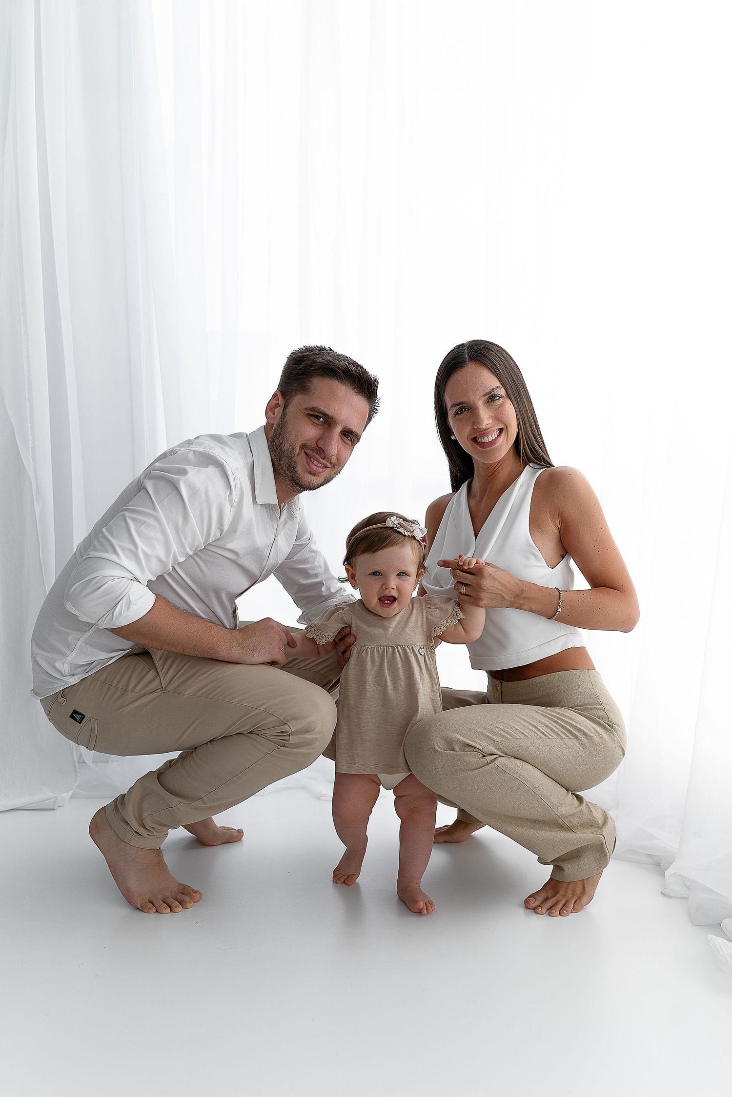A family in neutral clothing crouches together on white floor during studio photoshoot.