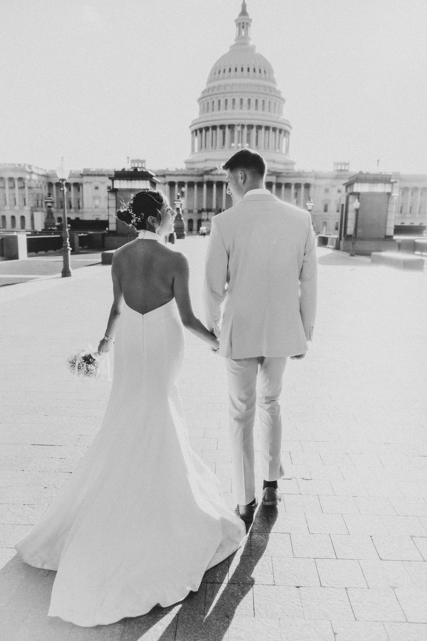 Black and white photo of couple in wedding attire walking toward US Capitol building.