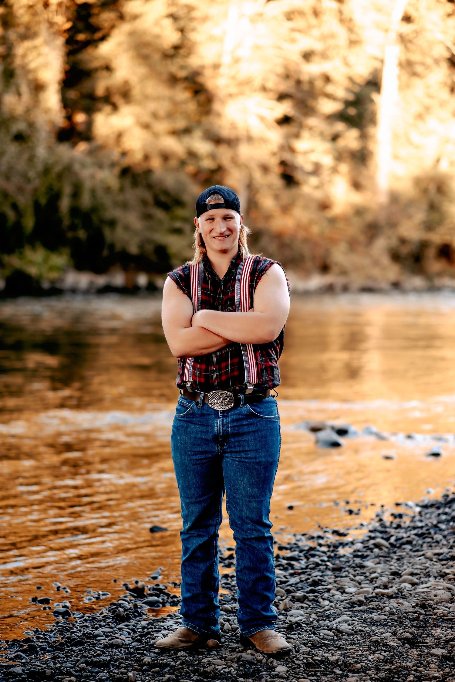 Person in jeans and vest sits casually by riverside during golden sunset.