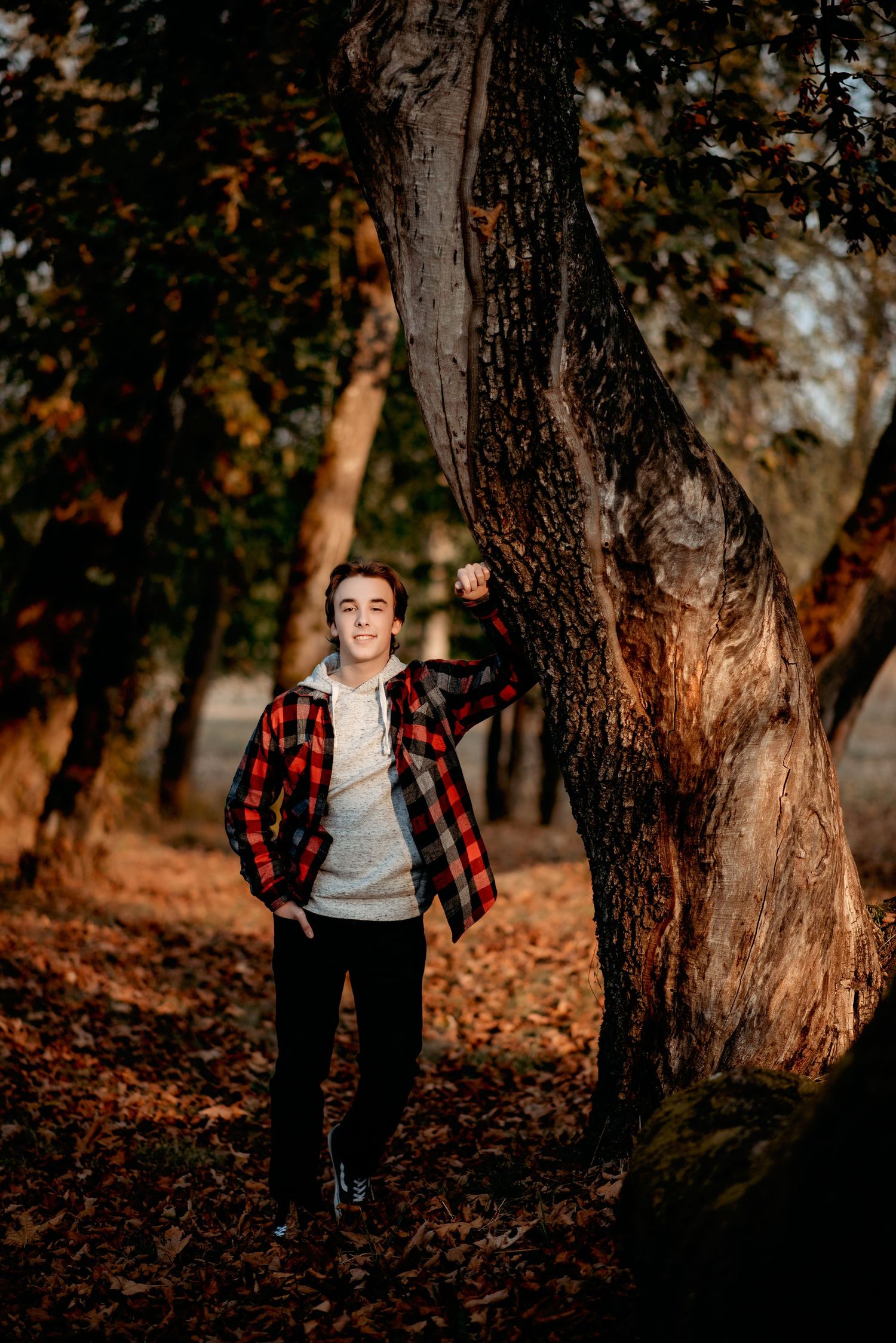 Person in buffalo plaid shirt poses among autumn trees at sunset.