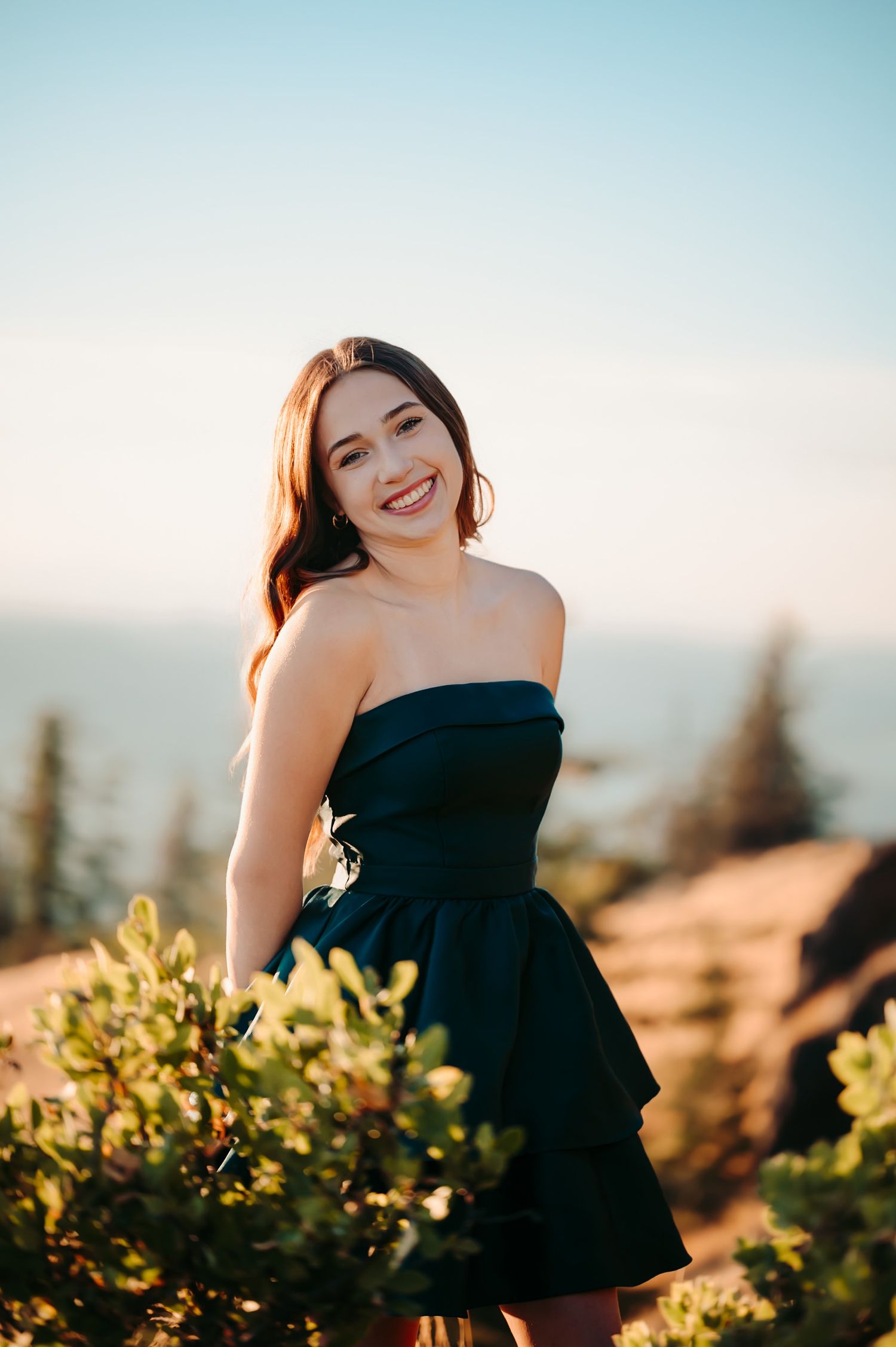 Person in black strapless dress poses among sunlit foliage at sunset.