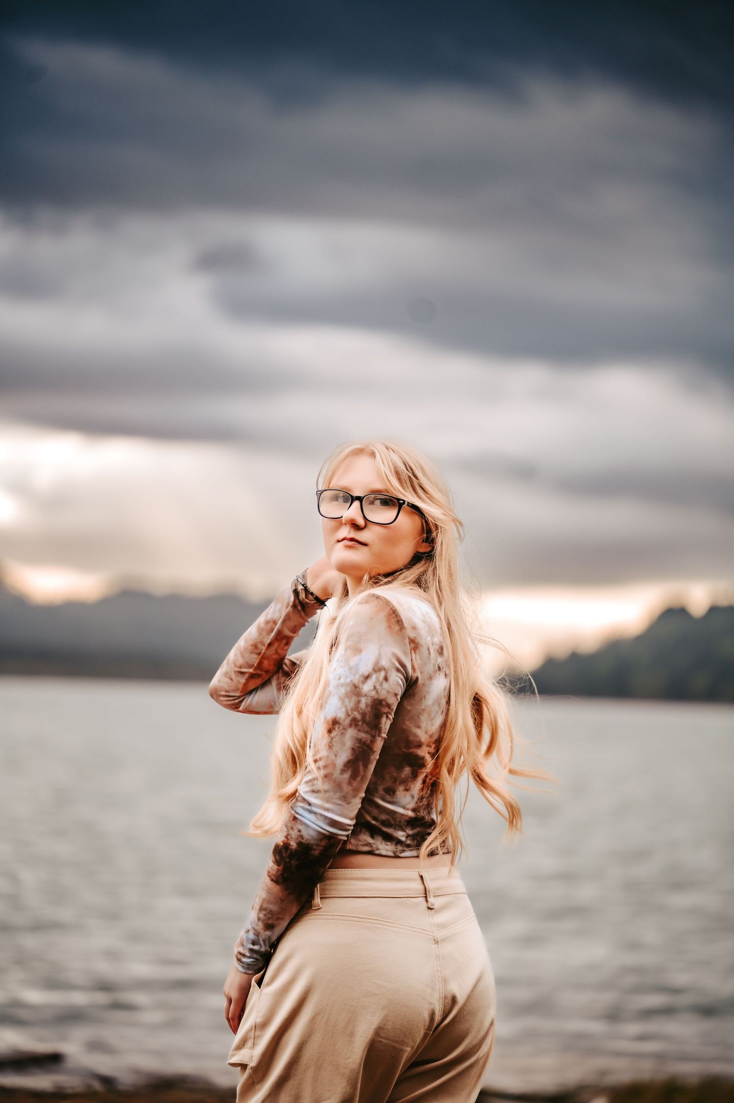 Someone in glasses poses by a lake during sunset with dramatic storm clouds in the background.