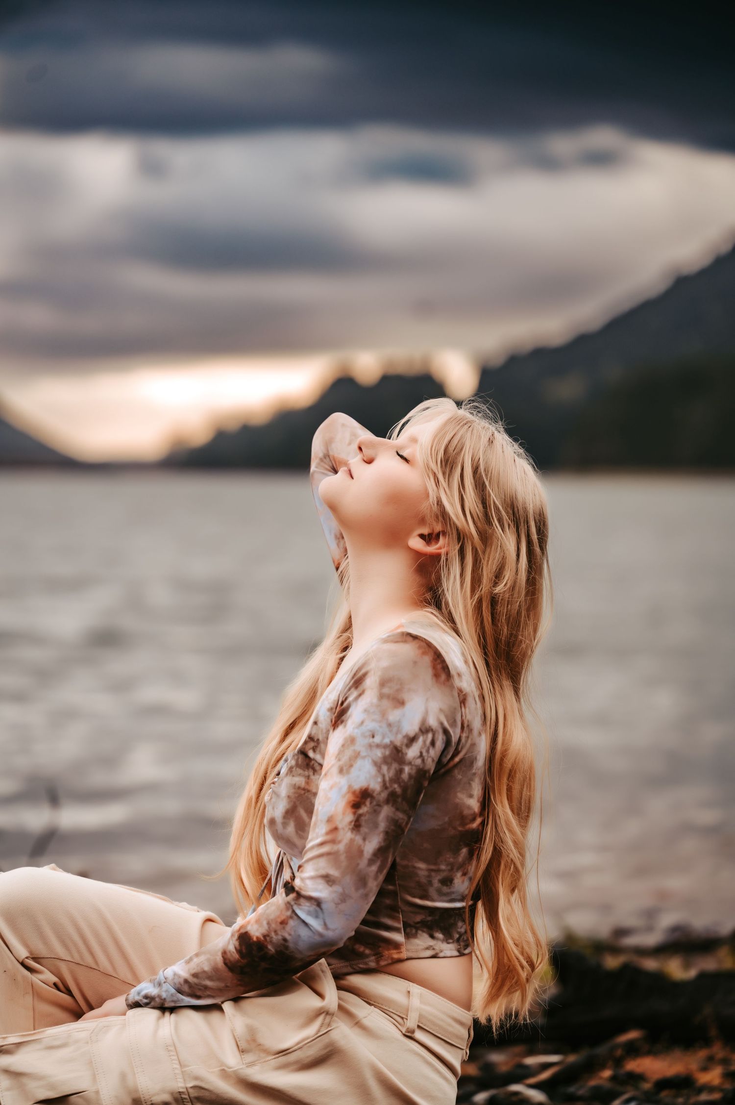 A dreamy outdoor portrait with someone looking up at dramatic cloudy skies near water.