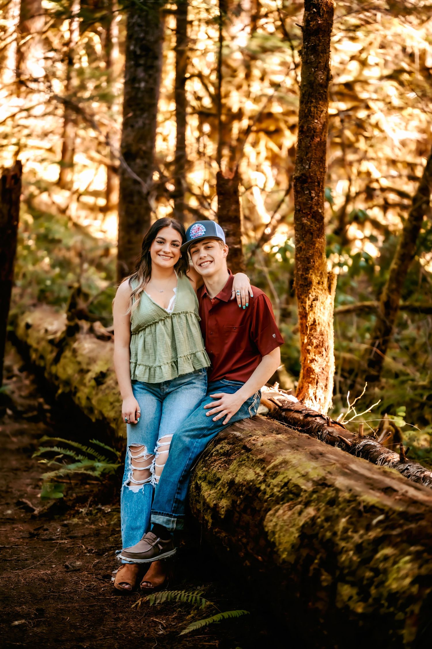 A couple sits together on a fallen log in a sunlit forest during golden hour.