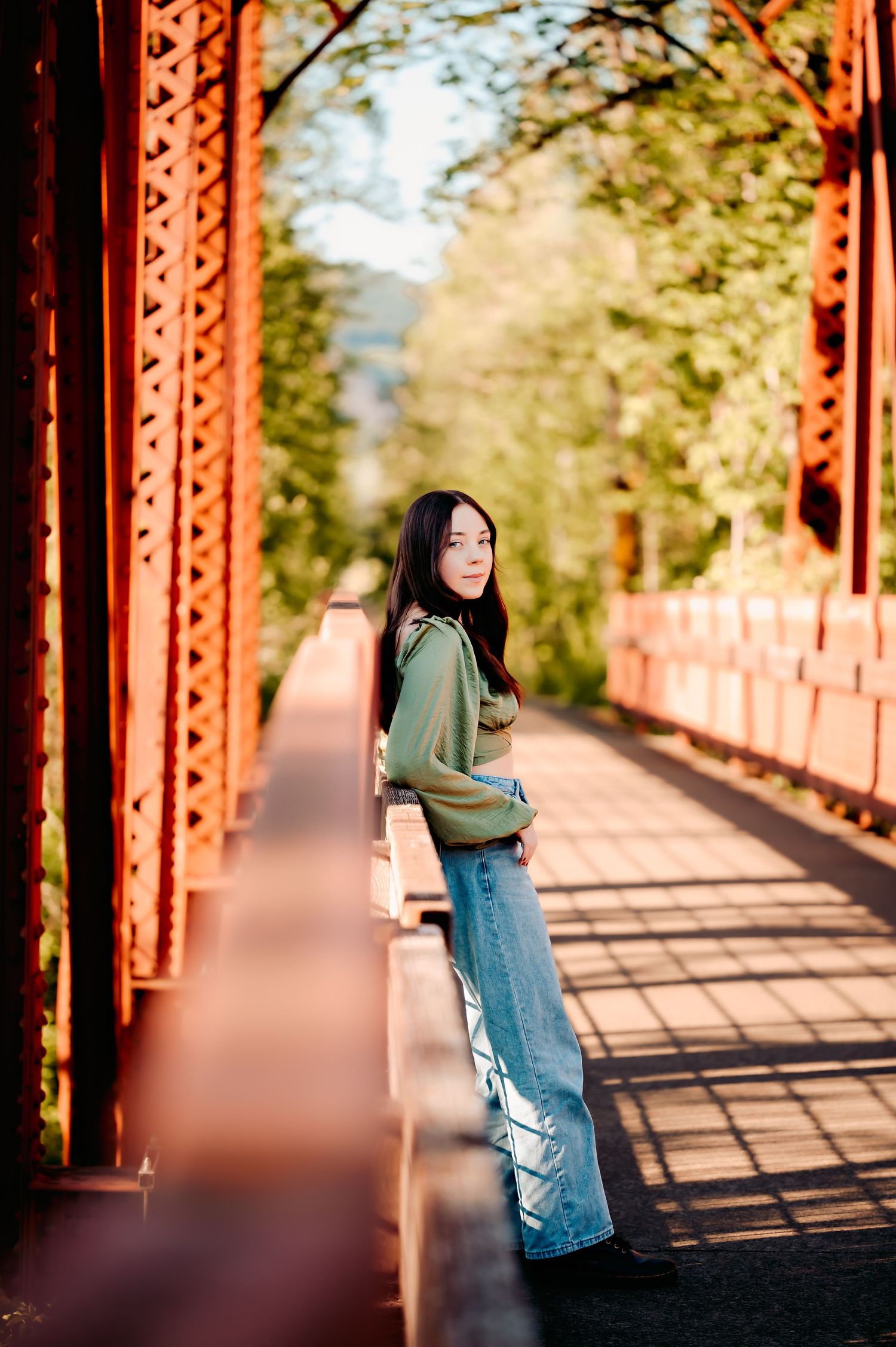 Person in denim and green top posing on red metal bridge during sunset.