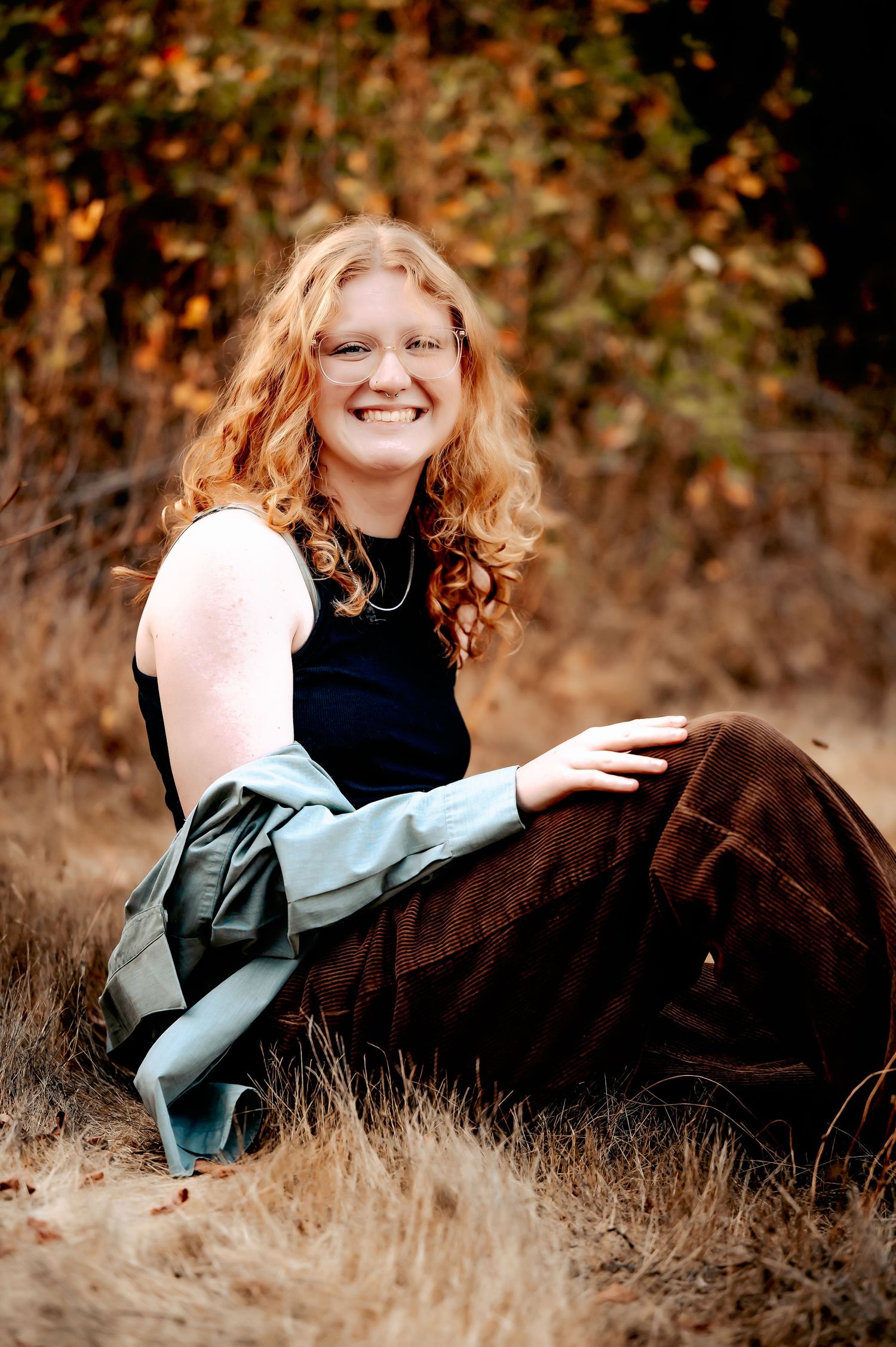 Someone in a dark dress sits in autumn grass with warm lighting and fall foliage in background.