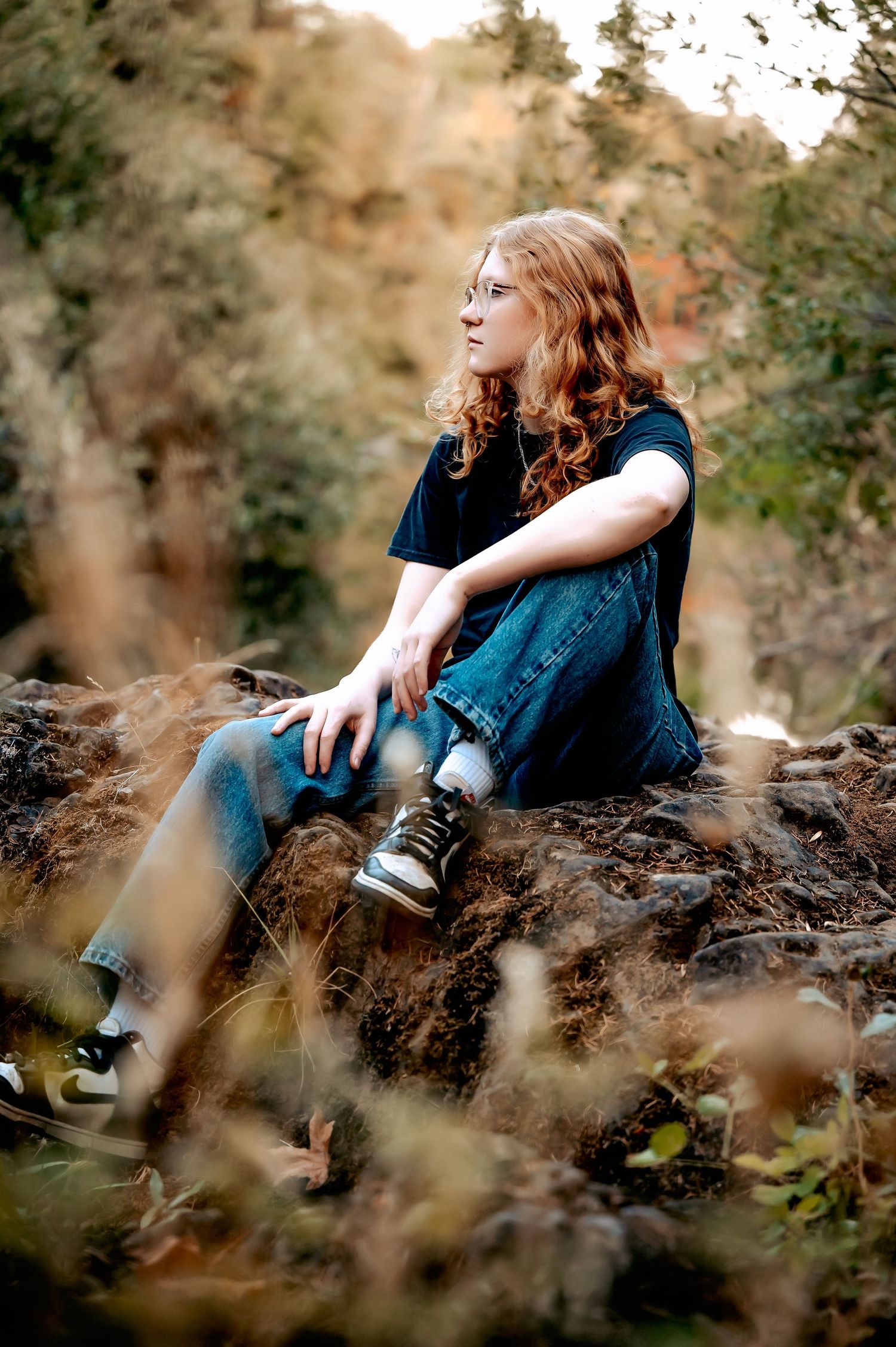 Someone in denim sits casually on rocky terrain with golden sunlight filtering through trees.