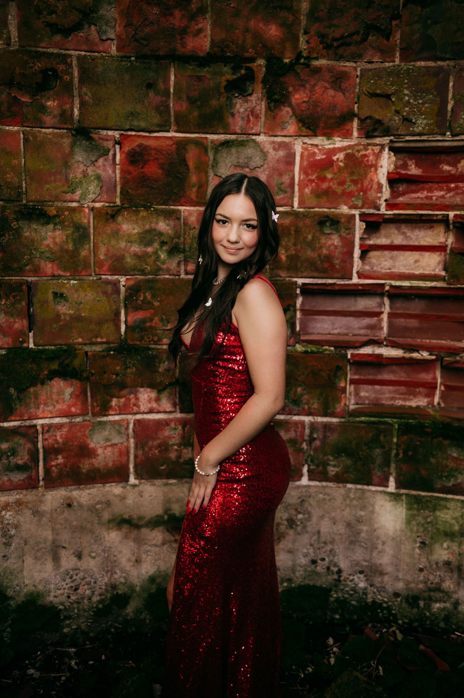 A person in a sparkly red evening dress poses against a rustic brick wall in dramatic lighting.