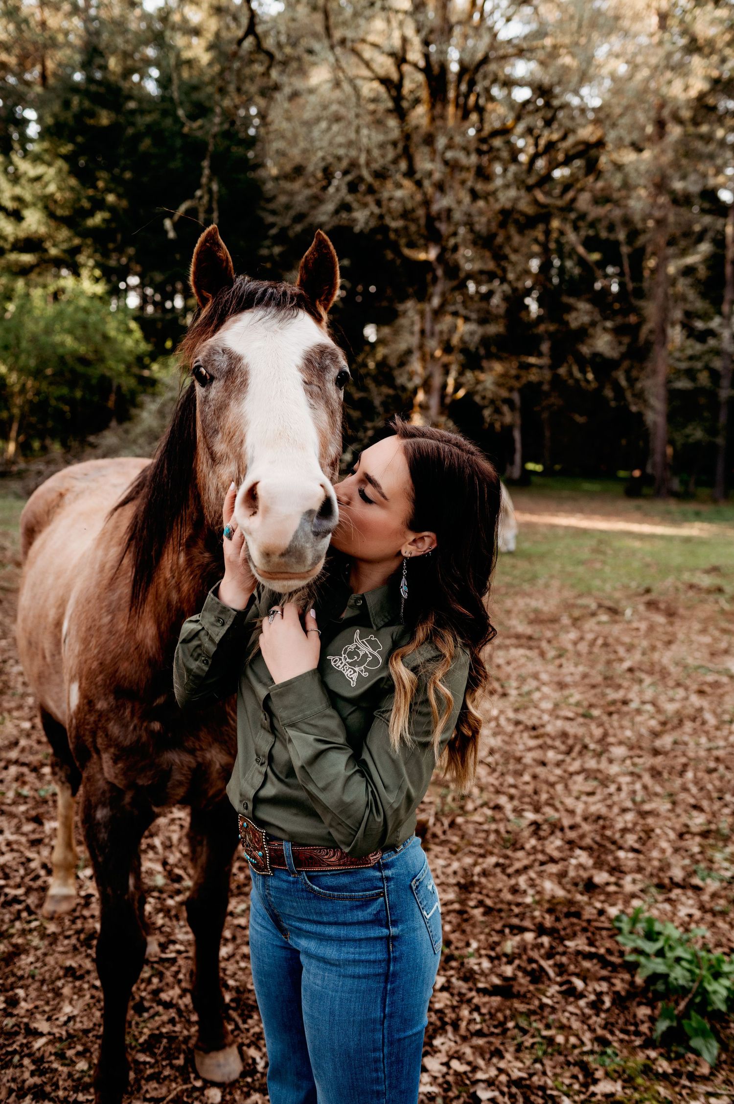 A brown and white horse receives affection from someone wearing a green jacket and jeans in an autumn forest setting.