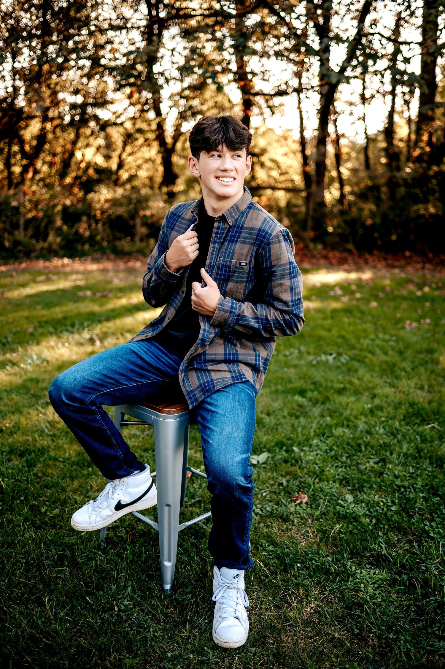 A person in a plaid shirt and jeans sits casually on a small metal stool outdoors during golden hour.