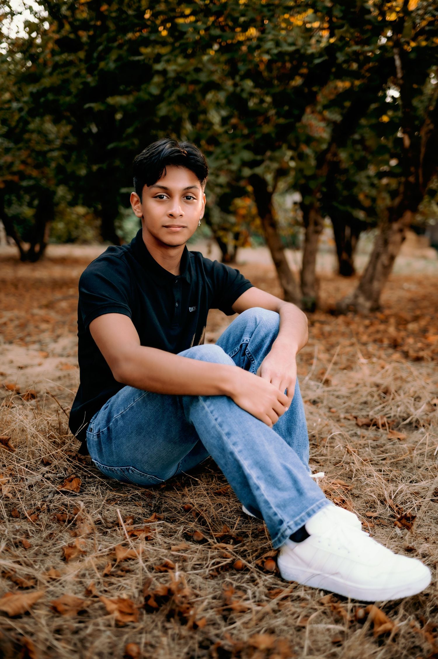 A person in a black shirt and jeans sits casually on fallen autumn leaves beneath trees in a park setting.