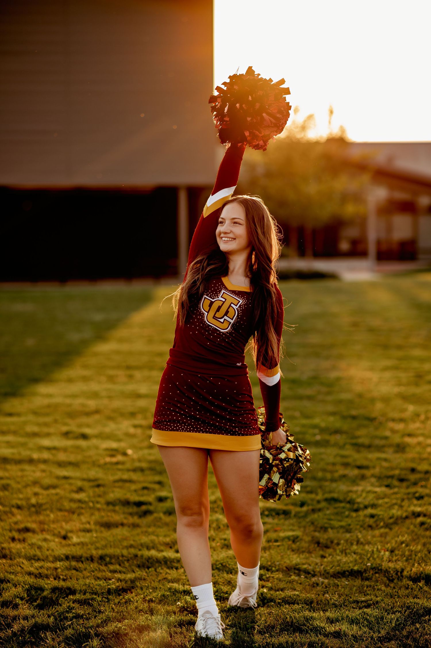 College cheerleader in maroon uniform raises pom pom during golden hour.