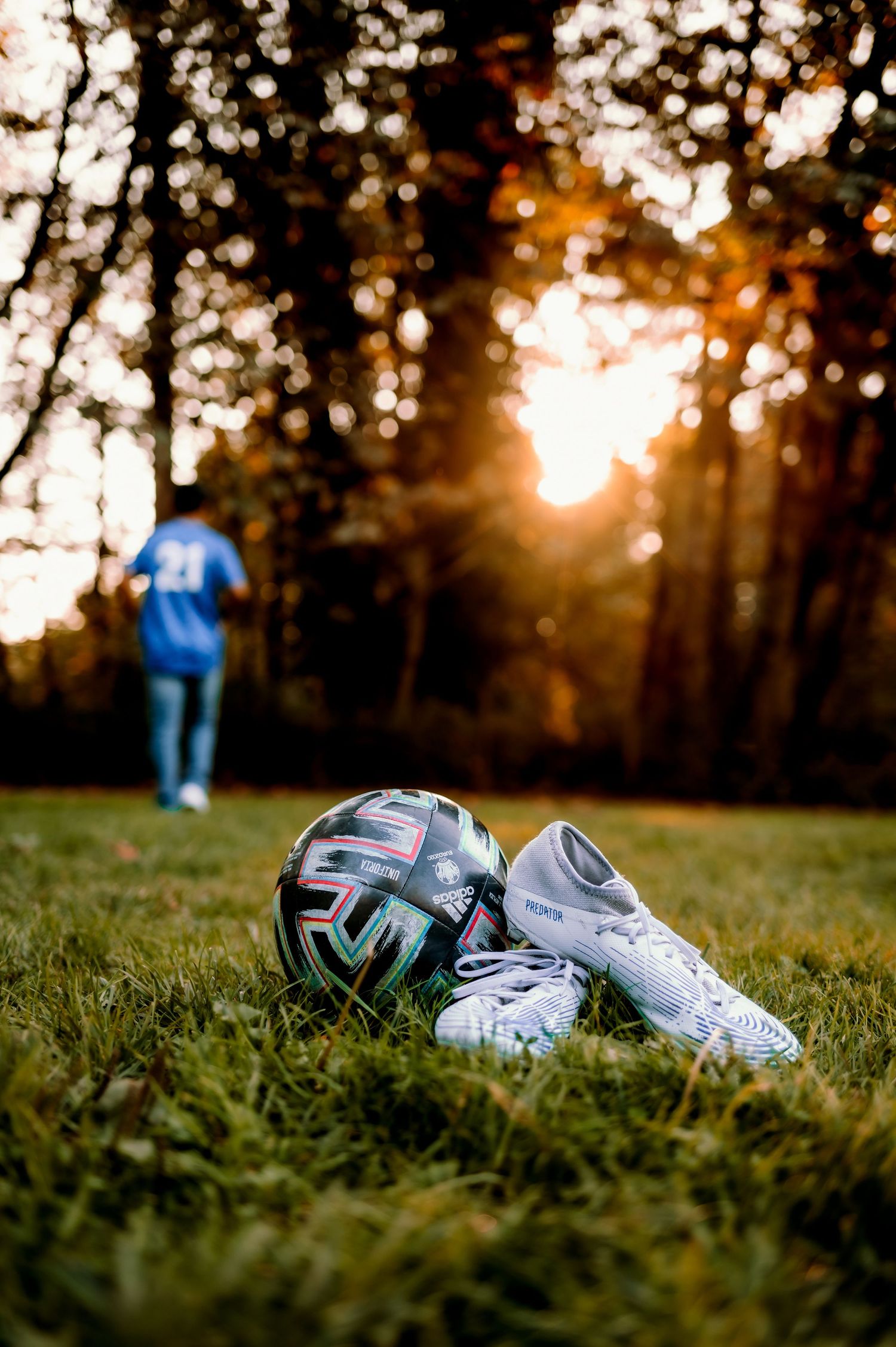 Soccer cleats lying on grass during golden hour with blurred figure in background.