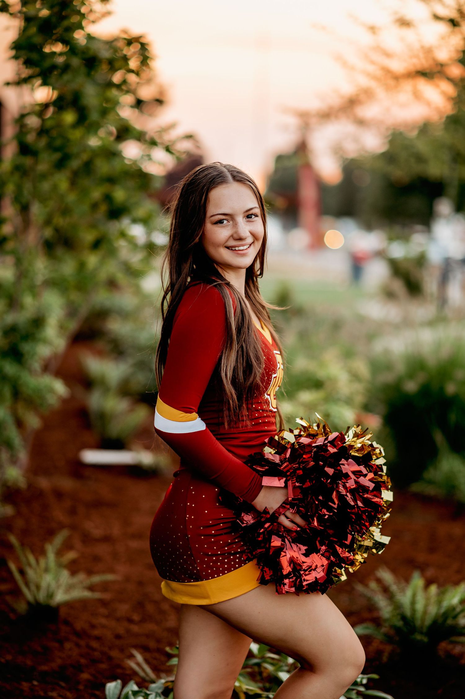 Cheerleader in red and gold uniform poses with pom poms at sunset.