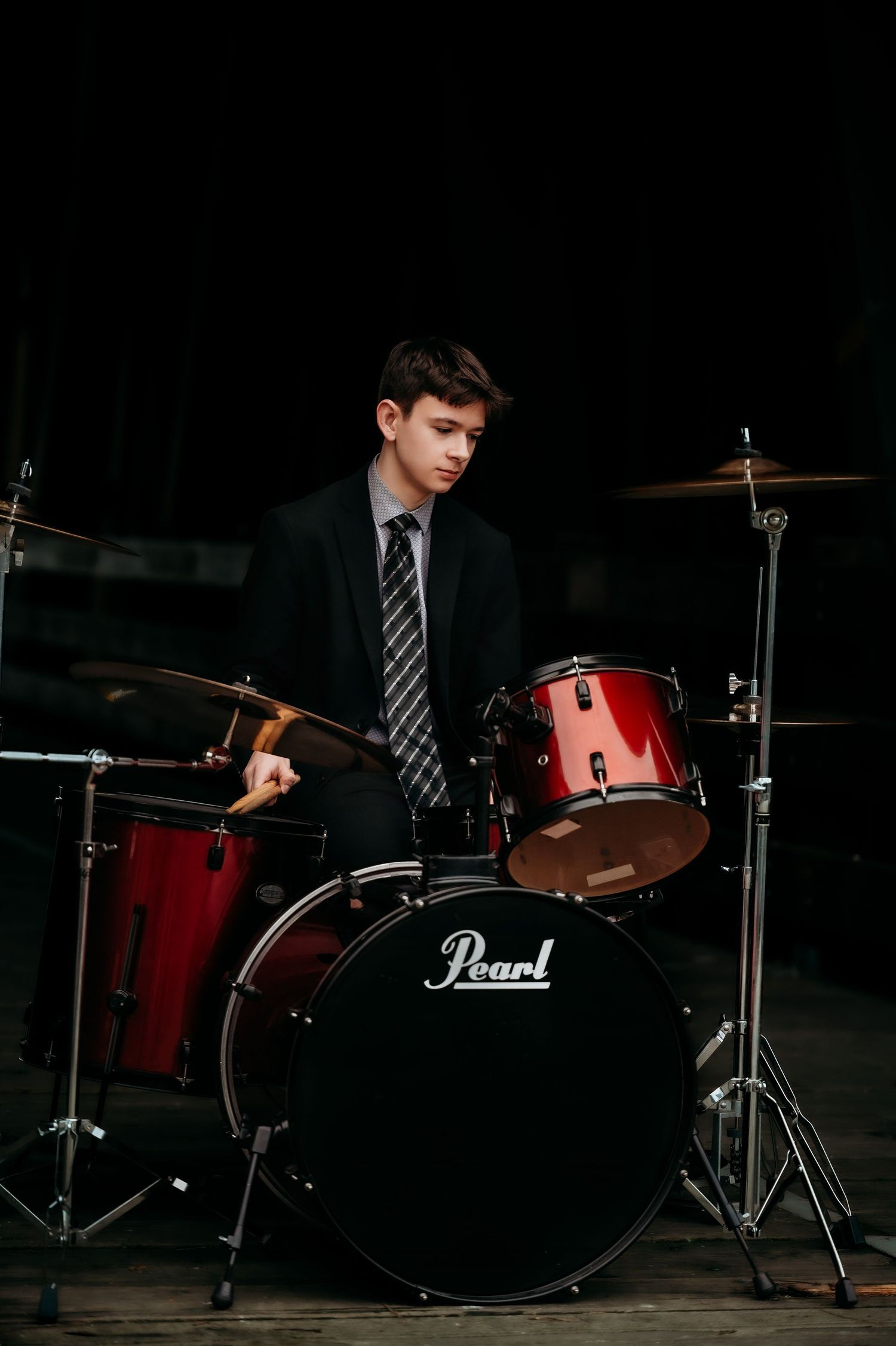 Musician in suit sitting at red Pearl drum kit against dark background.